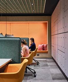 two women sitting at desks in an office setting with orange walls and grey flooring