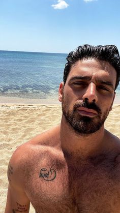 a man with no shirt on standing in front of the ocean and looking at the camera