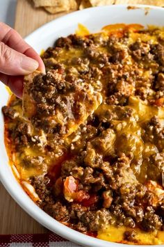 a hand holding a tortilla chip over a casserole dish with cheese and ground beef