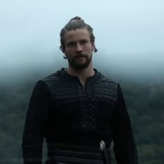 a man with long hair and braids standing in front of foggy trees wearing a black outfit