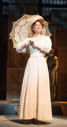 a woman in a white dress and hat holding an umbrella on stage with other people behind her