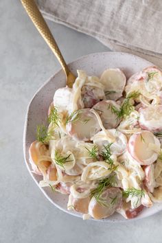 a white bowl filled with pasta and meat on top of a gray tablecloth next to a gold spoon