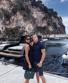 a man and woman posing for a photo in front of boats on the water at a marina