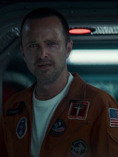 a man in an orange space suit looks at the camera while standing next to another man