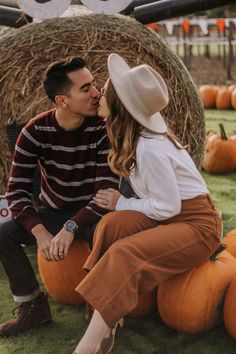 a man and woman sitting on pumpkins in front of hay bales, kissing