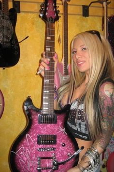 a woman holding a pink guitar in front of two guitars on the wall behind her