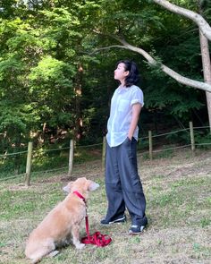 a woman standing next to a brown dog on top of a grass covered field with trees in the background