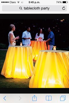 several people standing around yellow tablecloths in the grass with lights on them at night