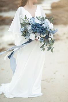 a woman in a white dress holding a blue and white flower bouquet on the beach