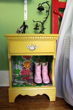 a yellow nightstand with pink boots and flowers on the top, in front of a green wall