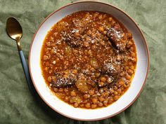 a white bowl filled with beans and meat on top of a green cloth next to a spoon