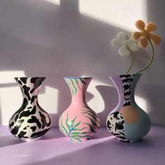 three vases with flowers in them on a purple tablecloth, one is painted pink and the other two are black and white