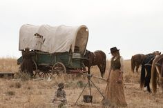 a woman in a dress and hat standing next to a horse drawn wagon on dry grass