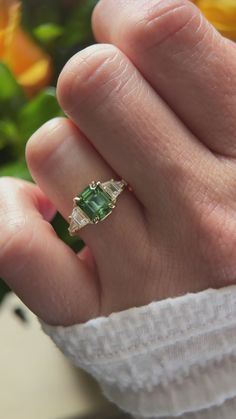 a person's hand holding a ring with a green stone in it and yellow flowers behind them