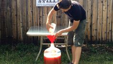 a man is pouring something into a jug on a table outside in the grass with a wooden fence behind him