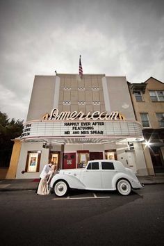 two people standing in front of a movie theater with an old car parked on the street