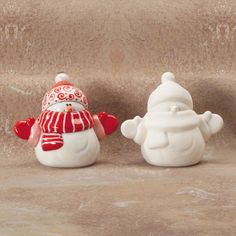 two small ceramic snowmen sitting next to each other on a stone surface, one wearing a red and white hat