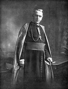 an old black and white photo of a man in a priest's robe, standing with his hands on his hips