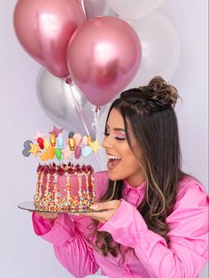 a woman in pink holding a cake with candles on it and some balloons behind her