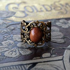 10x8mm brown goldstone cabochon on an antiqued brass adjustable filigree ring. Easy to adjust to any size. Shade of brownish orange may vary slightly if at all. About the stone: This is a man made gemstone. Thousands of tiny copper particles glitter against a brown background. The story has it that a monk accidentally dropped copper shavings into a vat of molten glass to create this material. Adjustable Bronze Rings With Antique Finish, Antique Adjustable Bronze Rings, Adjustable Oval Brown Rings, Adjustable Oval Rings In Brown, Vintage Adjustable Amber Ring, Vintage Adjustable Filigree Ring Gift, Adjustable Antique Brown Jewelry, Antique Adjustable Brown Jewelry, Brass Filigree