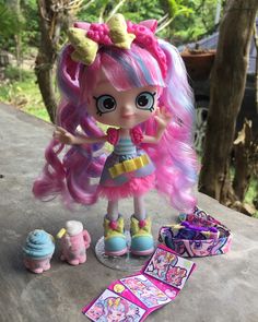 a doll with pink hair standing next to other dolls and accessories on a table in front of some trees