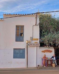 two people sitting at a table in front of a white building with an olive tree