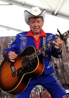 an old man with a cowboy hat and blue pants holding a guitar in his right hand