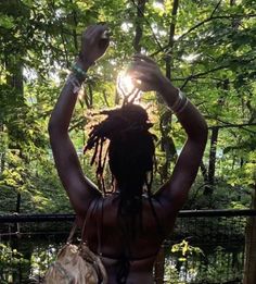 a woman with dreadlocks standing in front of trees and holding her hands up to the sky