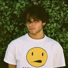 a man wearing a t - shirt with a smiley face on it, standing in front of some bushes