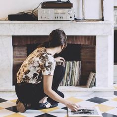 a woman kneeling down on the floor looking at a book in front of a fireplace