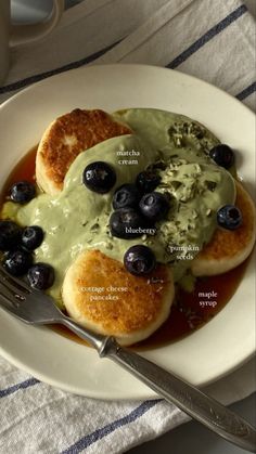 a white plate topped with pancakes covered in green sauce and blueberries next to a cup of coffee