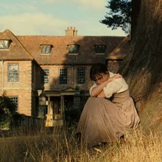 a woman sitting on the ground next to a large tree in front of a house