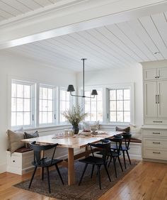 a dining room table with chairs and a bench in front of two windows on the wall