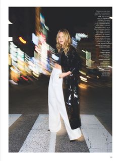 a woman in black jacket and white pants crossing street at night with city lights behind her