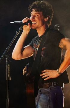 a man singing into a microphone while holding a guitar in his right hand and wearing a black shirt