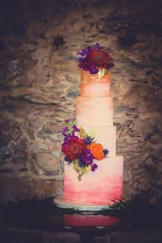 a multi - tiered cake with flowers on top sits in front of a stone wall