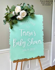 a welcome sign for a baby shower with white flowers on it and greenery in the corner