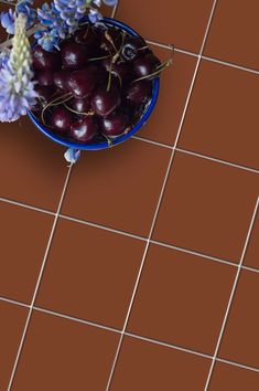 a blue bowl filled with cherries sitting on top of a tiled floor next to purple flowers