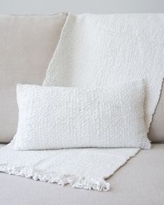 two white pillows on a couch with a blanket over the back and one pillow that has fringed edges