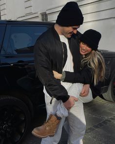 a man and woman walking in front of a black car with their arms around each other