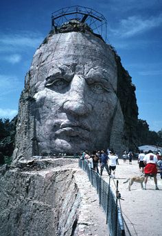 a large head on the side of a mountain next to a fence and people walking around