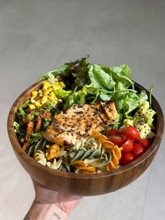 a hand holding a wooden bowl filled with pasta, vegetables and chicken on top of it