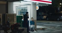 two people sitting on the curb in front of a gas station