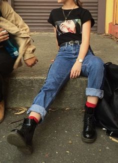 a woman sitting on the curb with her legs crossed and wearing black boots, jeans and a t - shirt