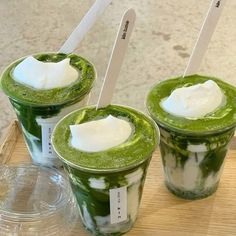 three green smoothies with whipped cream in them on a wooden tray next to a plastic cup