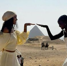 two women in white dresses are pointing at each other with pyramids in the background