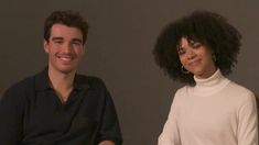 two people sitting next to each other in front of a gray wall smiling at the camera