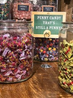 several glass jars filled with candy on top of a wooden table next to a sign that says, penny candy that's still a penny