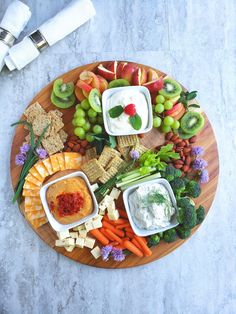 a wooden platter filled with fruit and veggies next to crackers, dip