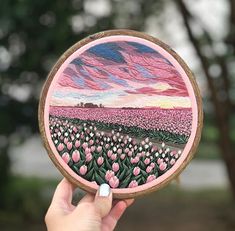 a person holding up a painted wooden plate with pink flowers on the front and green trees in the background